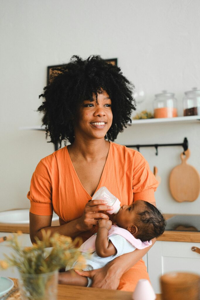 A mother in an orange dress smiles while feeding her baby with a bottle in a stylish kitchen.
